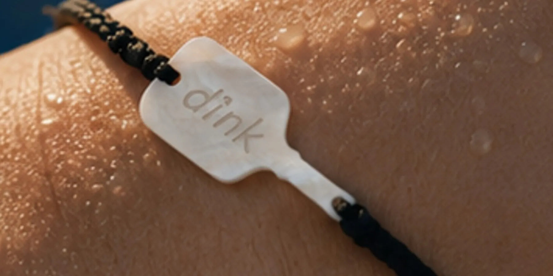 a black bracelet with a stone paddle charm with the word "dink" on a sweaty wrist