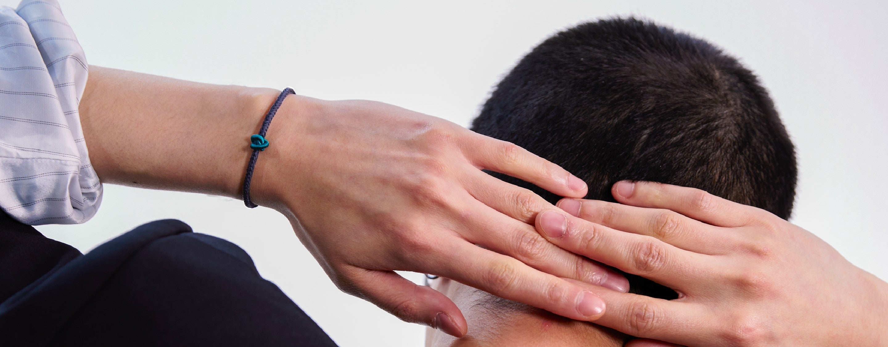 Person with hands wearing a green knot bracelet on his person's head, possibly in a comforting gesture, against a plain background.