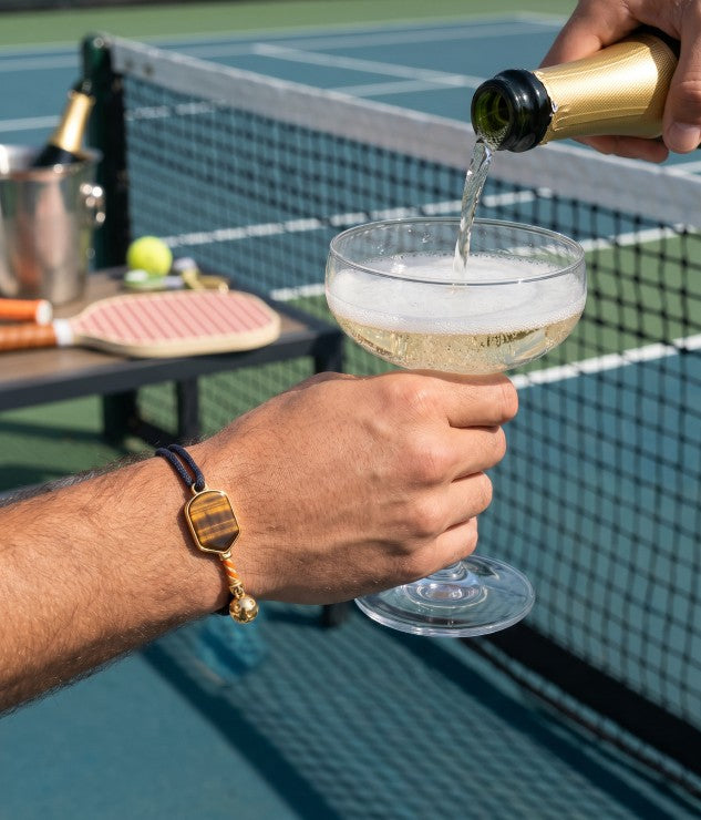 Person pouring champagne into a glass on a tennis court and wearing Glint & Luck's pickleball bracelet