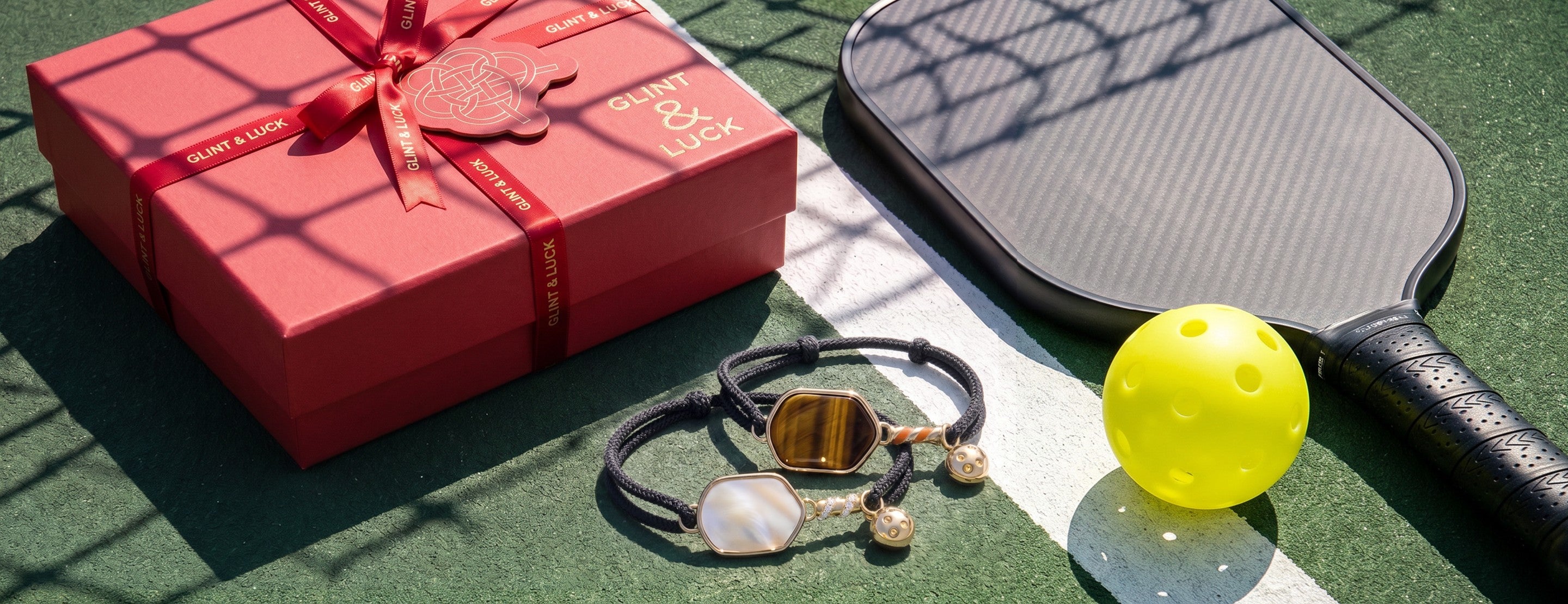 Red gift box, pickleball paddle, Glint & Luck black and white pickleball bracelets, and yellow ball on a green surface