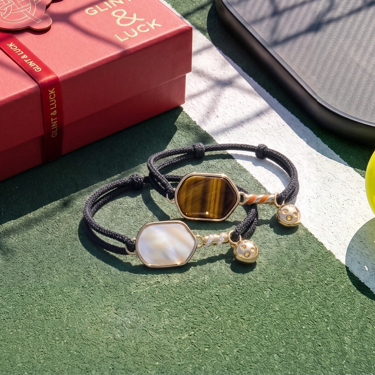 Two pickleball bracelets with paddle charms on a green surface next to a red box labeled 'Glint & Luck'.