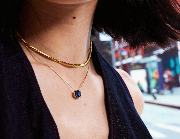 Close-up of a person wearing a gold necklace with a blue gemstone in an urban setting