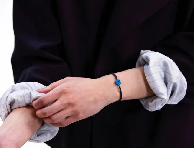 Person wearing a black cord bracelet with a blue knot on a plain background