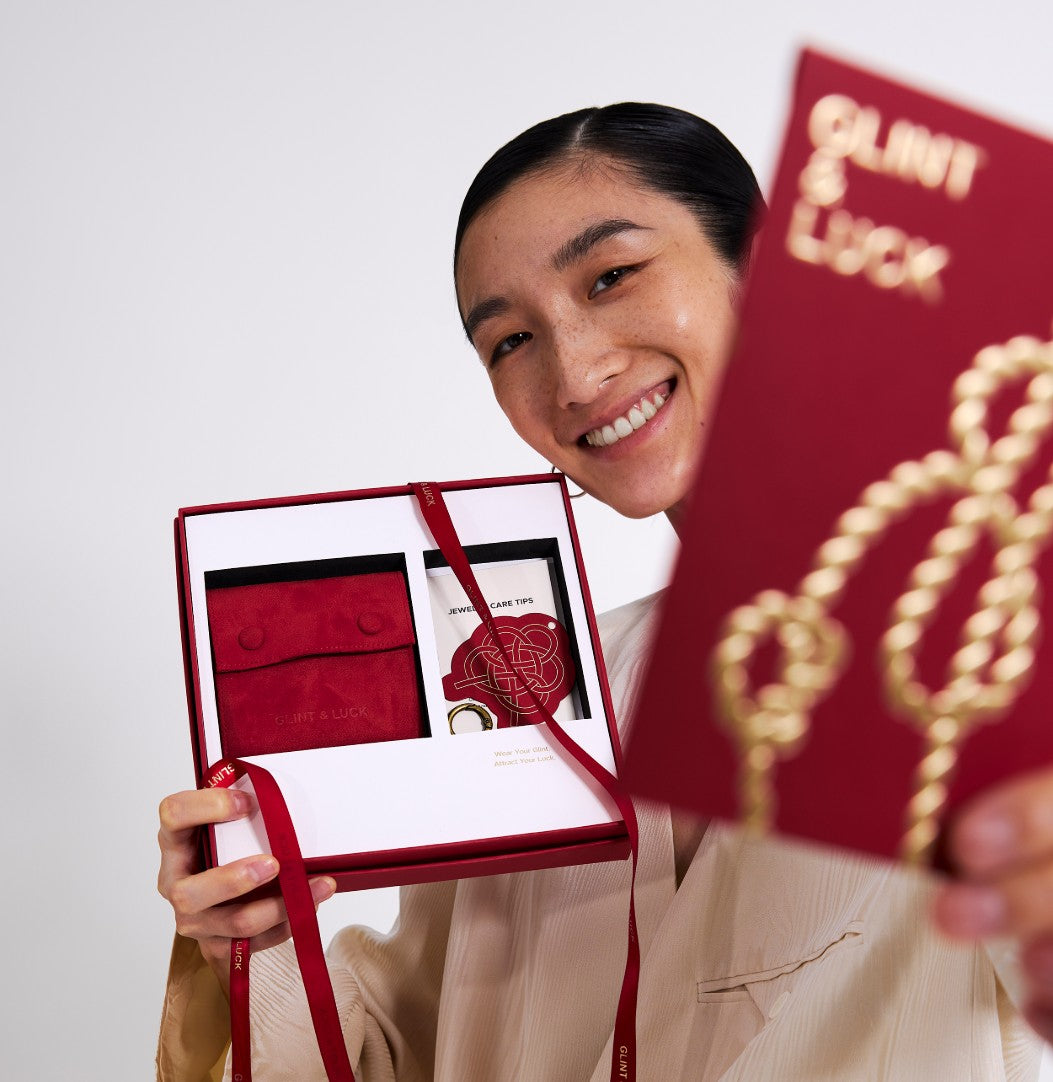 Person holding Glint & Luck red jewelry box and a red card with gold text against a white background