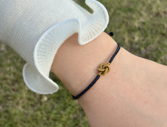 Close-up of a wrist wearing a black cord bracelet with a gold knot charm against a grassy background.