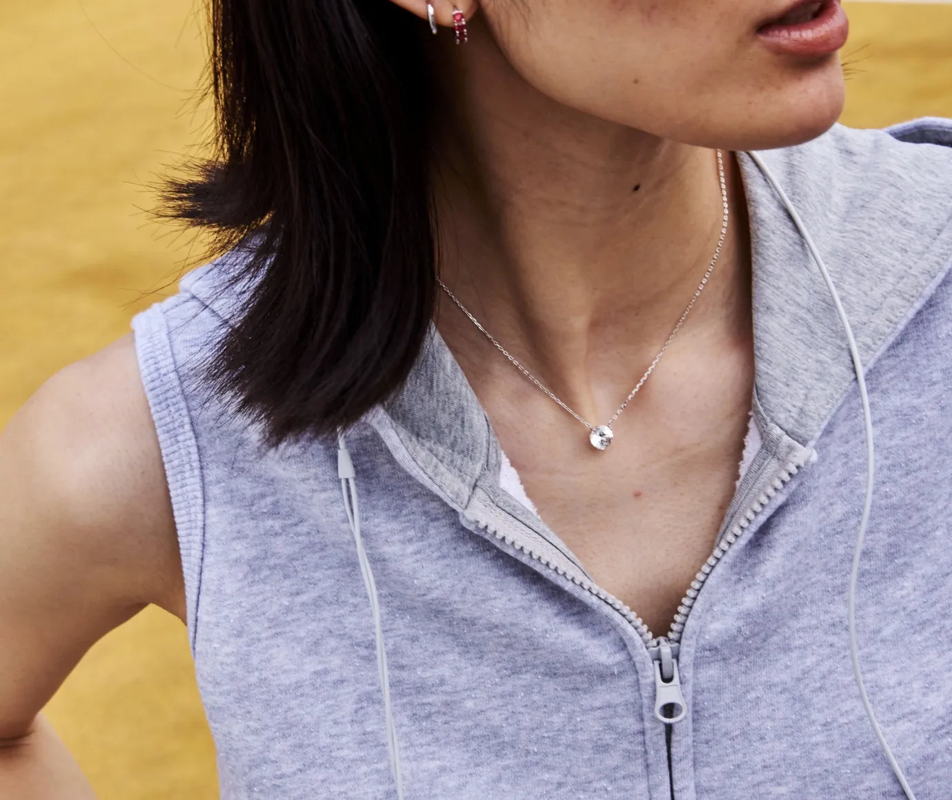Person wearing a silver necklace with white stone and a light gray sleeveless hoodie with a zipper, against a yellow background