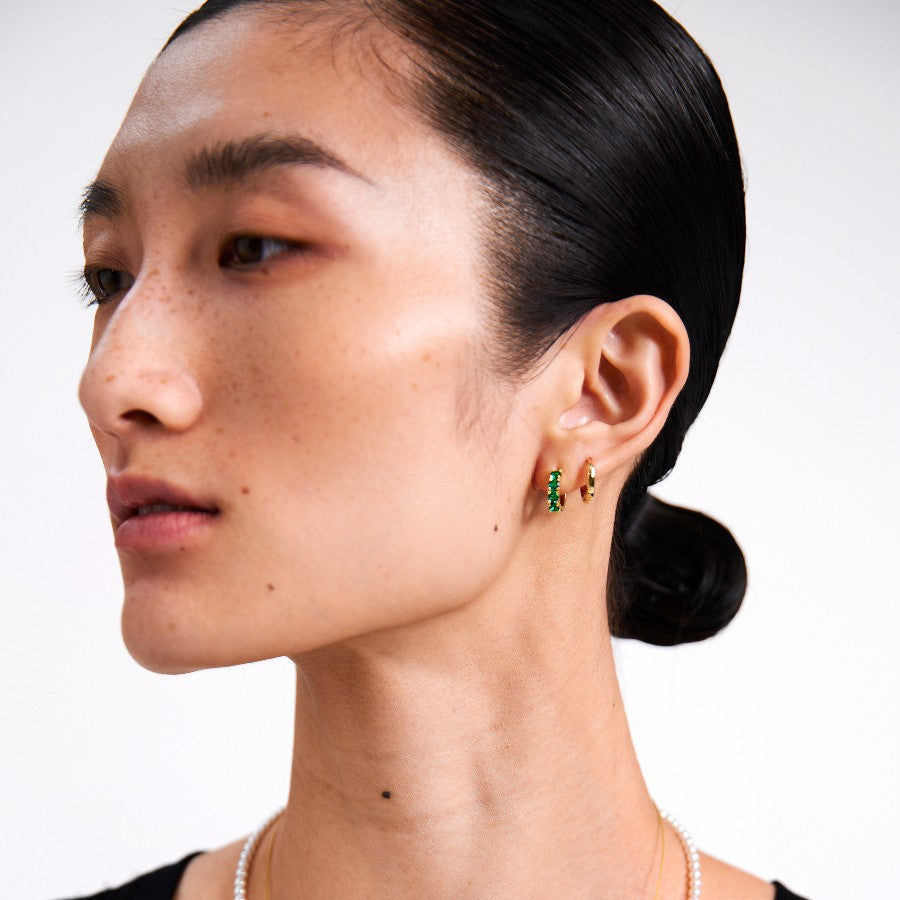 Close-up of a woman wearing emerald earrings and gold hoops with a plain background