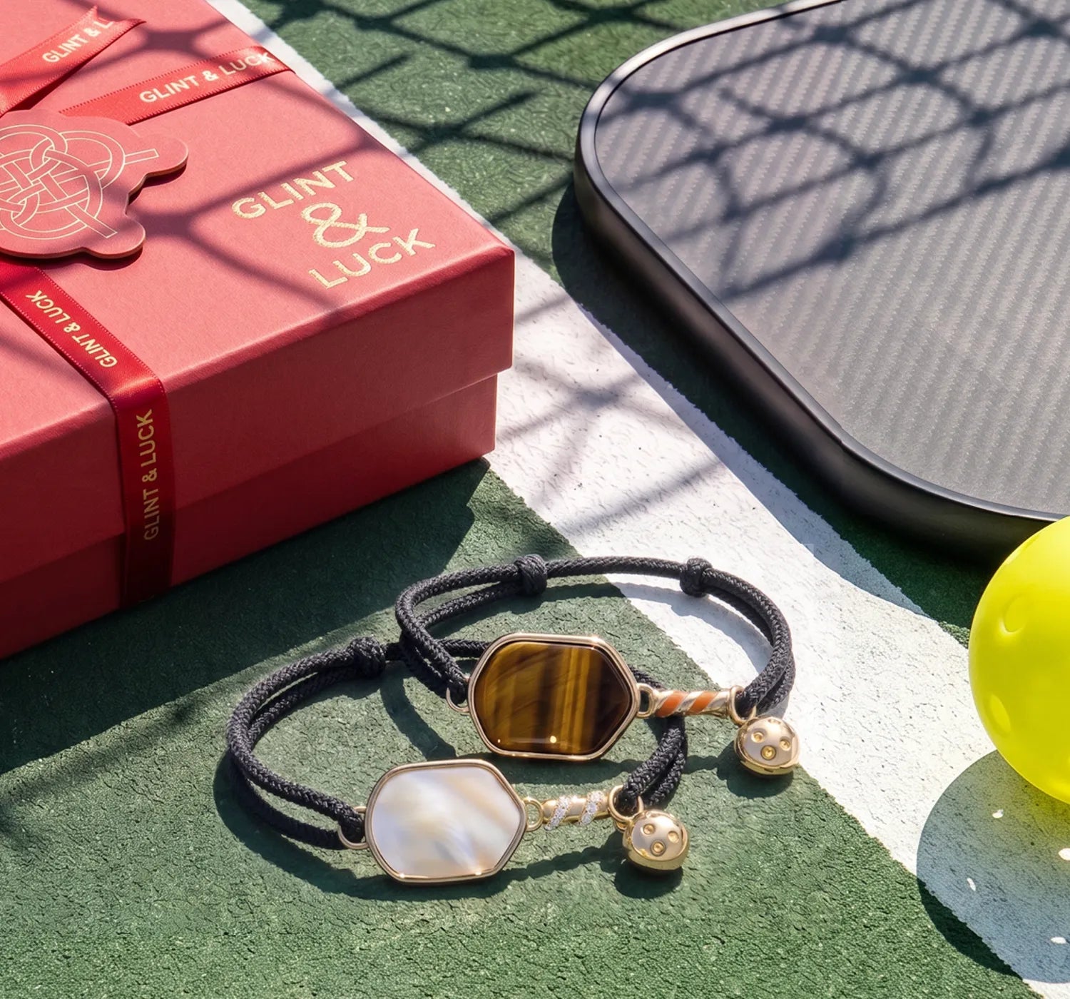 Two pickleball bracelets with decorative stones on a green surface next to a red box and yellow ball.