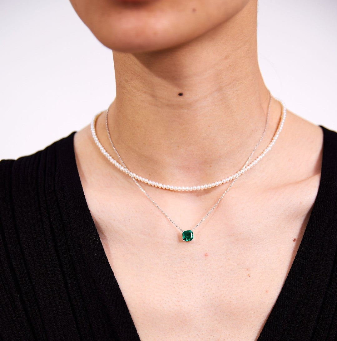 A pearl necklace, a silver necklace with a green gemstone pendant worn by a person on a plain background