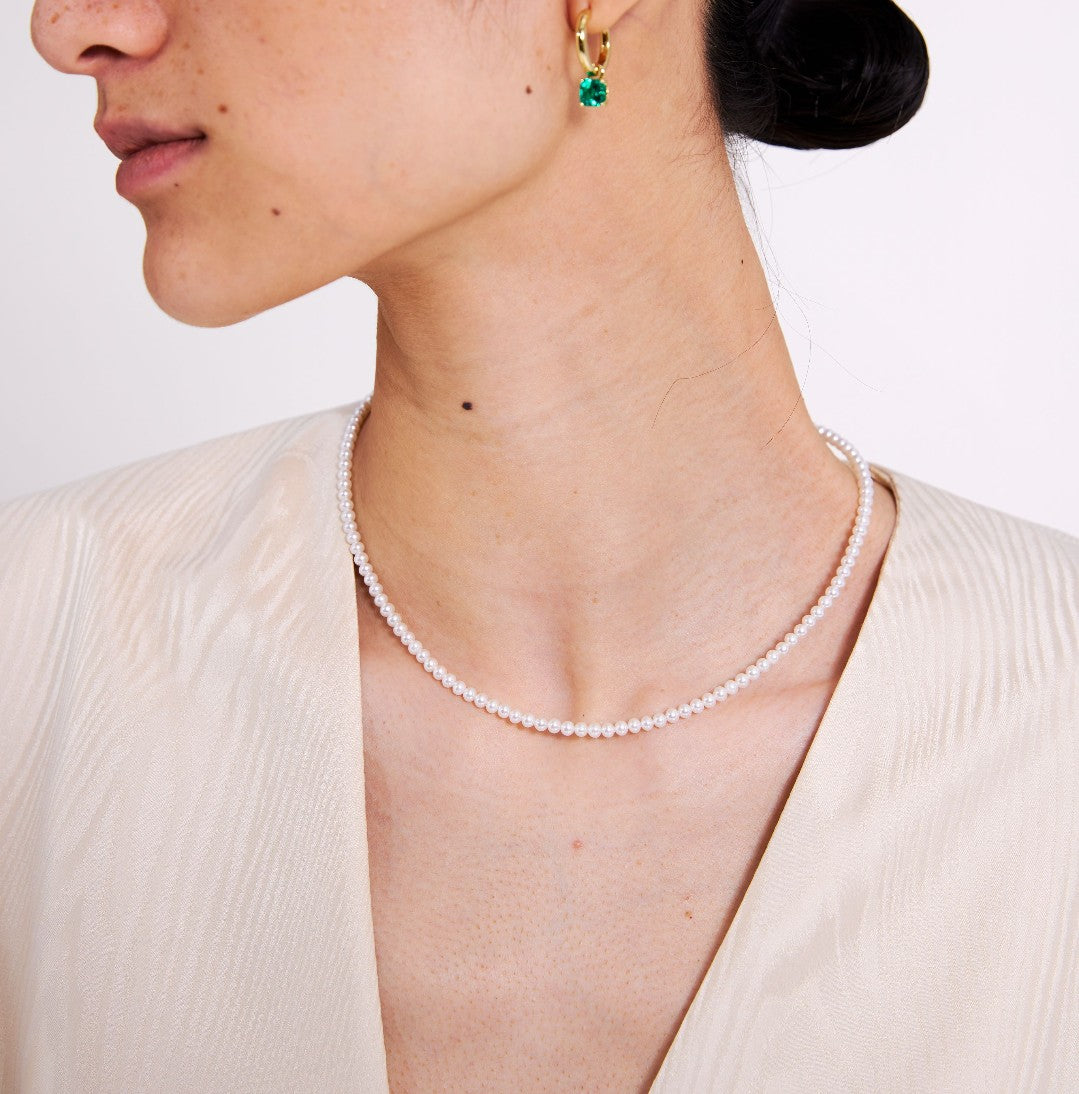 Close-up of a person wearing a pearl necklace and gold earrings with a green gemstone on a plain background