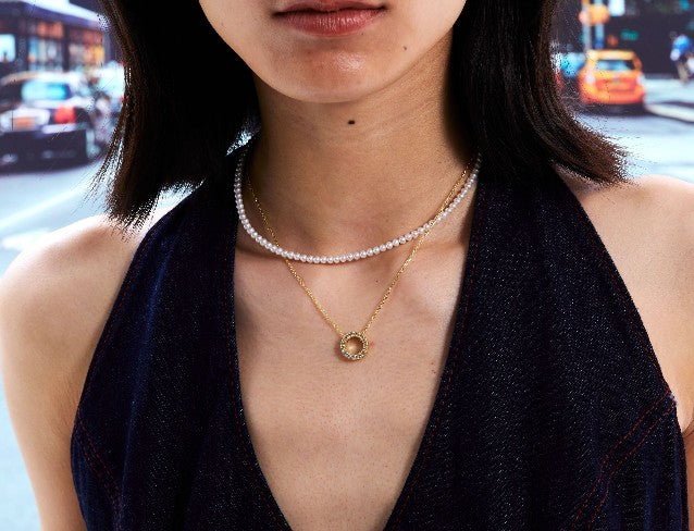 Close-up of a person wearing a pearl necklace and a gold necklace with round pendant with a blurred city street background