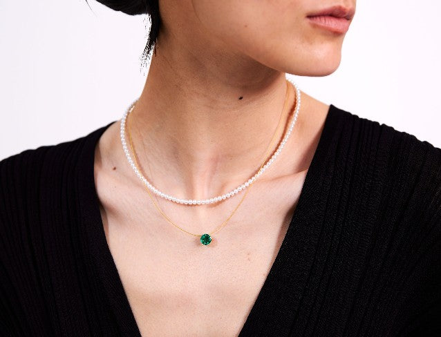 Person wearing a pearl necklace and wire necklace with a green pendant on a plain background