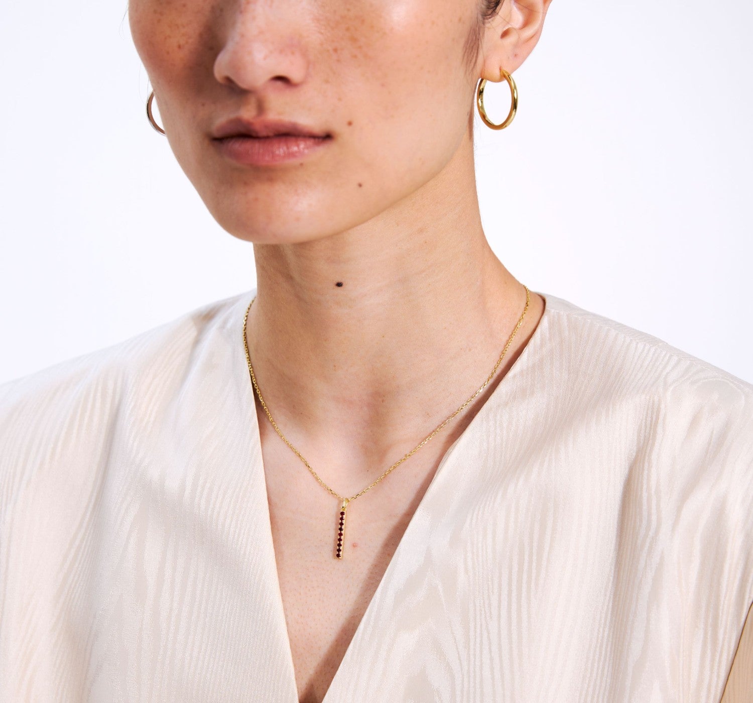 Close-up of a person wearing gold hoop earrings and a delicate gold bar necklace on a white background
